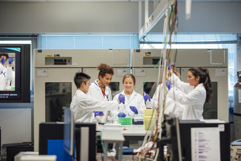 A group of science students stand in a group in a laboratory, they are being instructed how to run human samples. An older mixed race female teacher is encouraging them in their hands on practical lesson. The students stand around a central table. Various pieces of medical research equipment and supporting computer apparatus, layer the image, to create depth.The individuals seem positive. Gritty women.